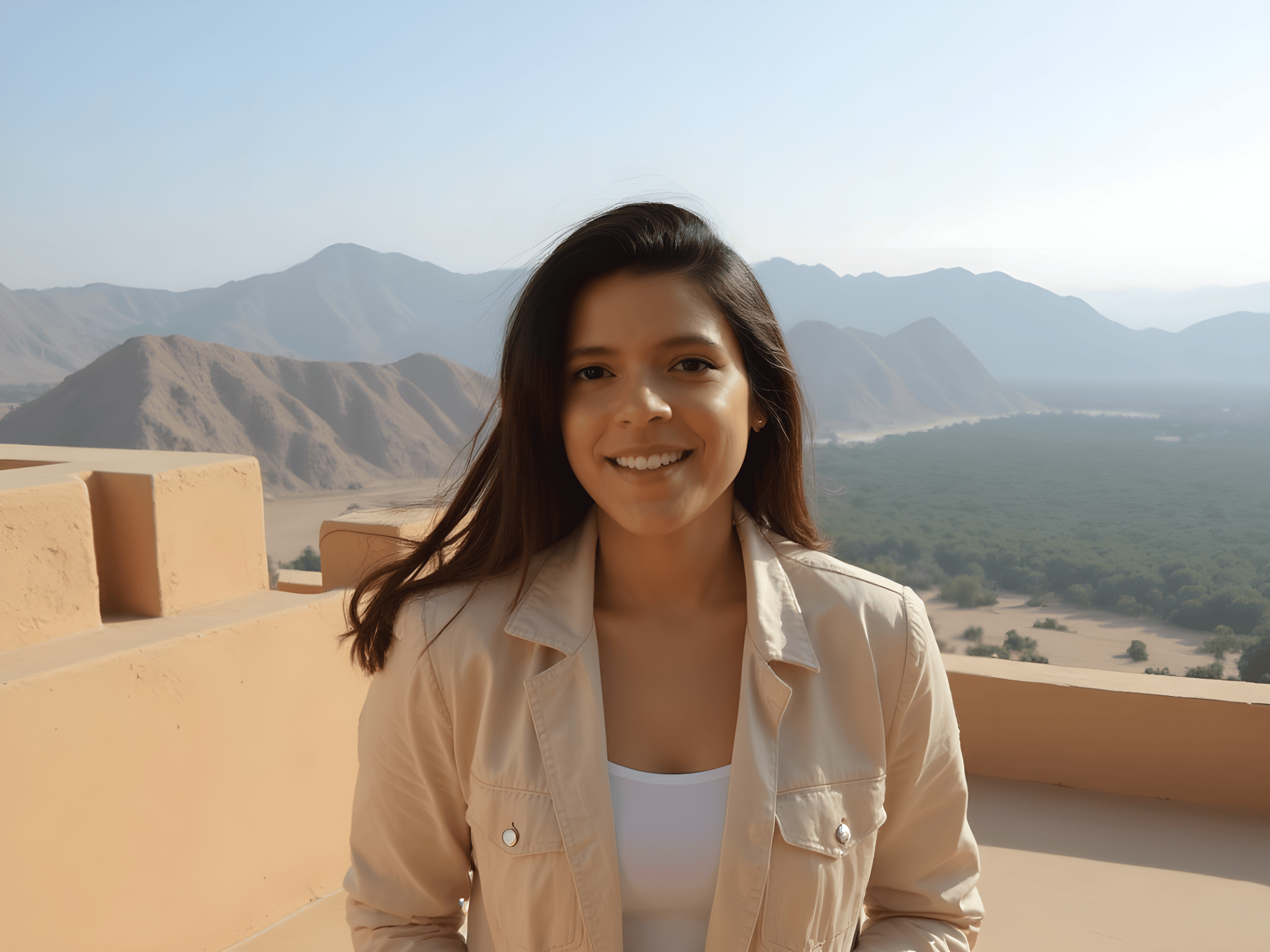 Smiling woman in a light jacket stands in front of scenic mountains and valley views.